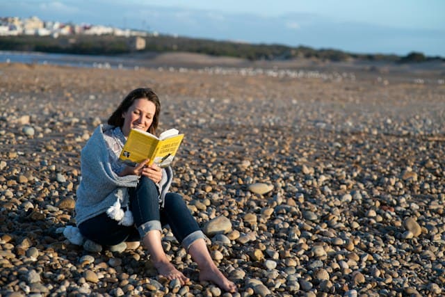 A woman sits contentedly by the rocky shores of a serene lake in New Zealand, absorbed in a book.
